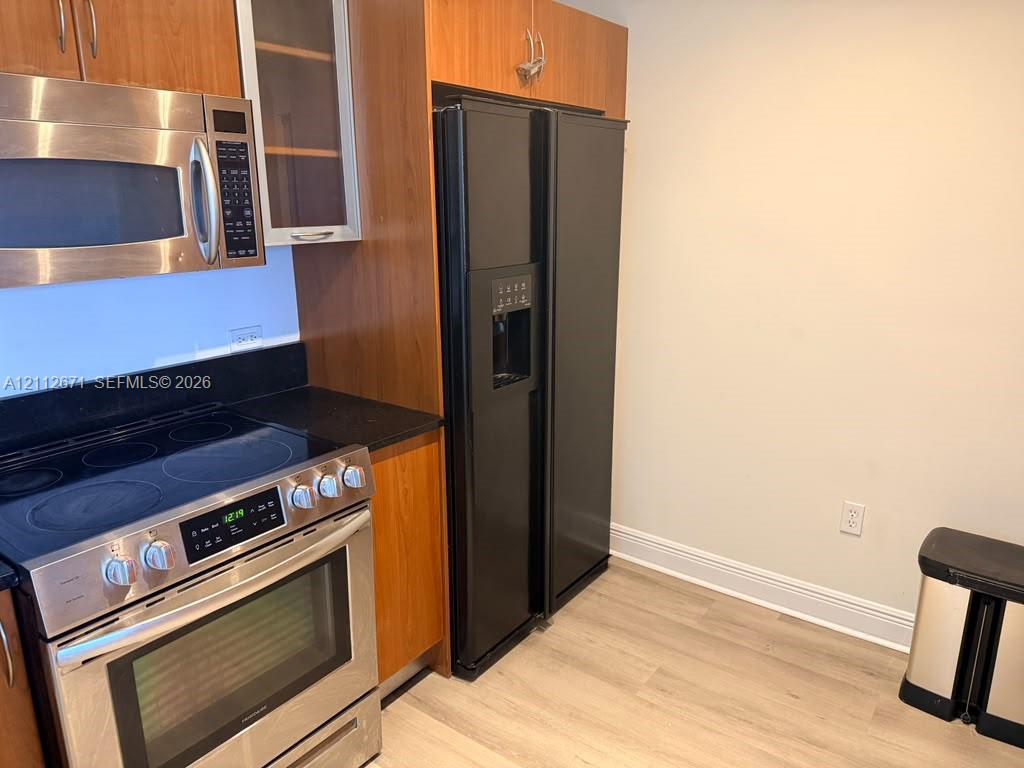 Kitchen featuring wood-finish cabinetry, stainless steel electric range, built-in stainless steel microwave, black side-by-side refrigerator, and dark solid-surface countertops