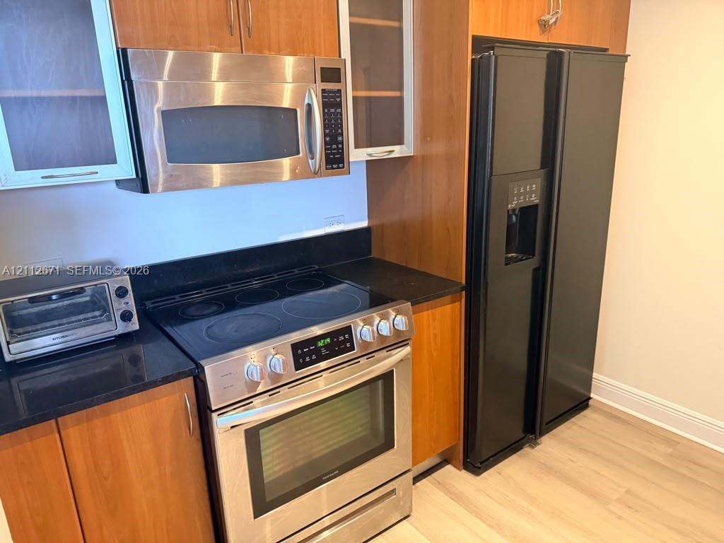 Kitchen featuring wood cabinetry, black solid-surface countertops, stainless steel microwave, stainless steel electric range, and a black refrigerator Kitchen featuring wood cabinetry, black solid-surface countertops, stainless steel microwave, stainless steel electric range, and a black refrigerator