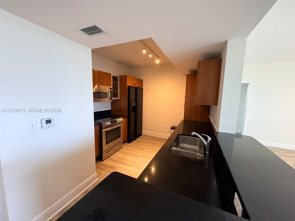 Kitchen featuring dark stone countertops, wood-finish cabinetry, stainless steel appliances, and track lighting Kitchen featuring dark stone countertops, wood-finish cabinetry, stainless steel appliances, and track lighting