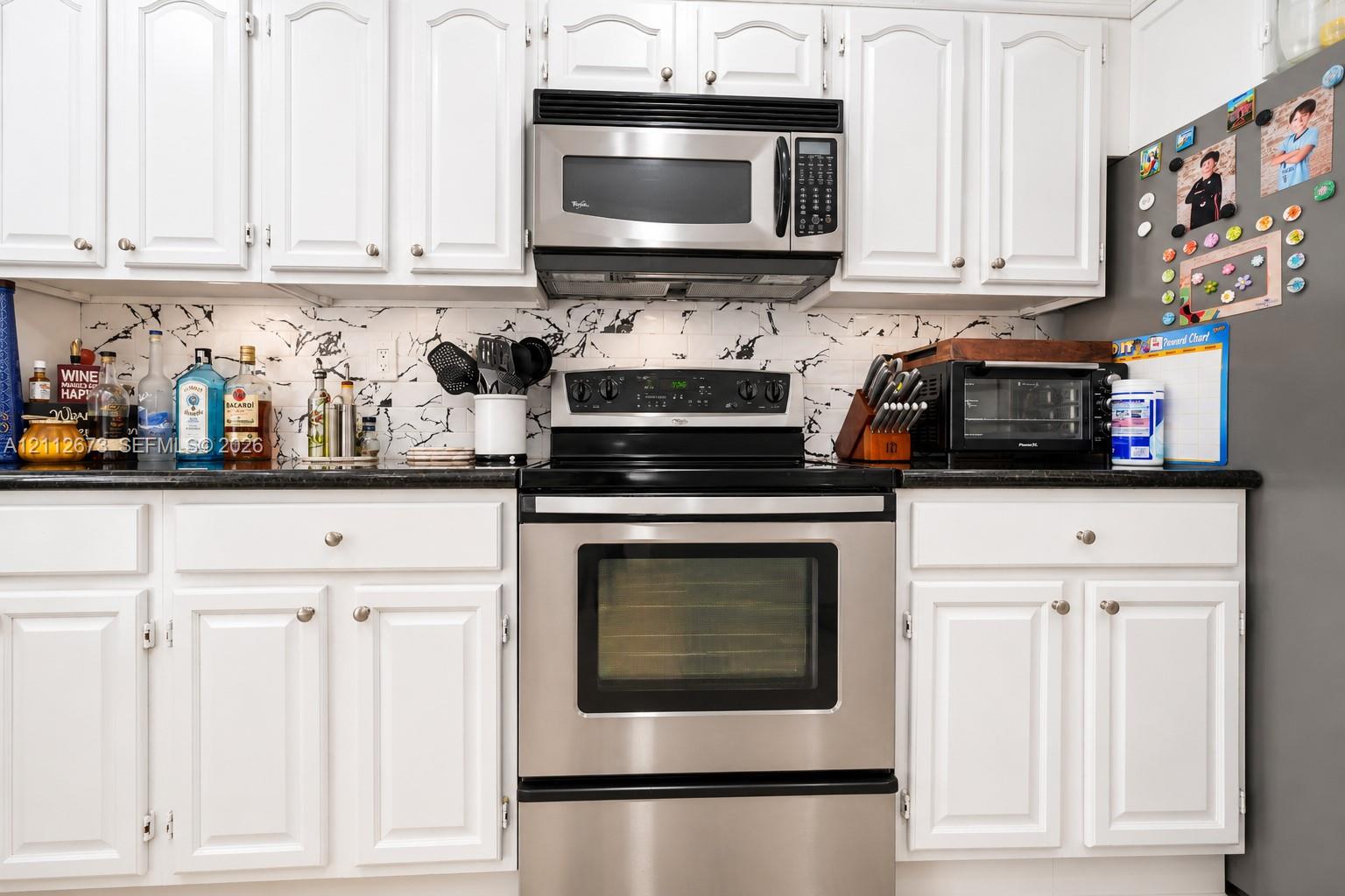 Kitchen featuring white raised-panel cabinetry, dark solid-surface countertops, patterned tile backsplash, and stainless steel appliances including an over-the-range microwave and range