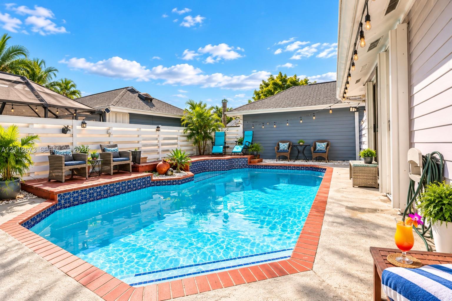 Outdoor swimming pool featuring decorative tile accents, a brick paver border, concrete patio, and white horizontal privacy fencing Outdoor swimming pool featuring decorative tile accents, a brick paver border, concrete patio, and white horizontal privacy fencing