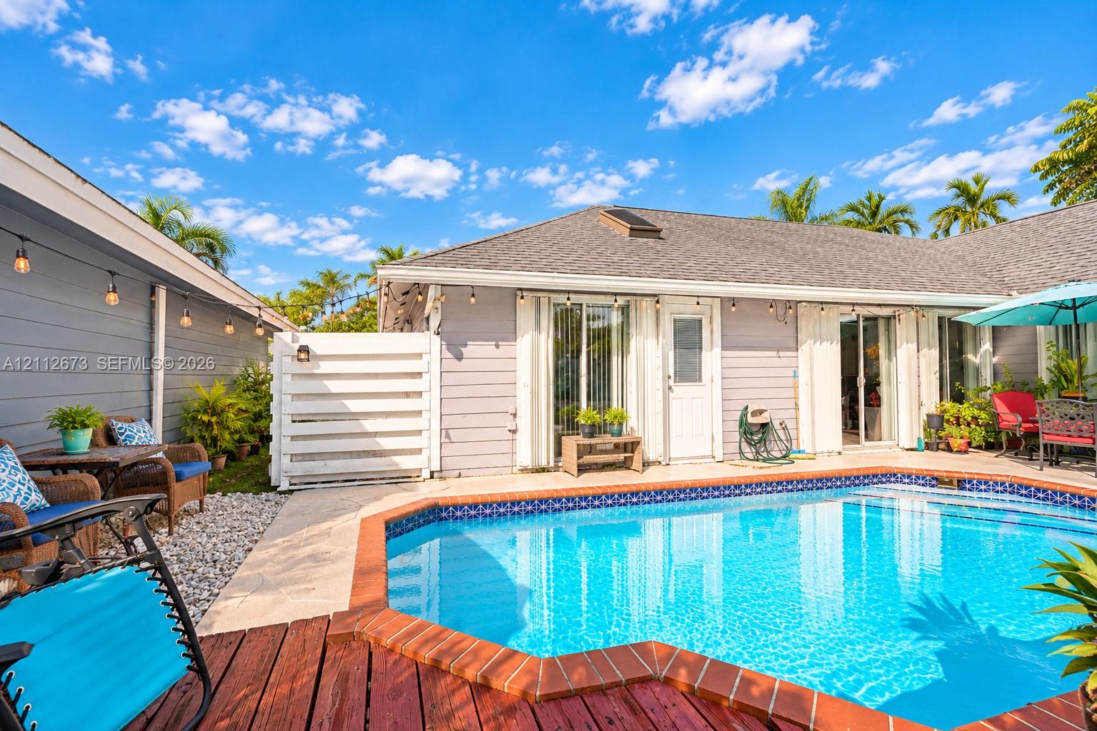Outdoor living area featuring a custom-shaped swimming pool with brick coping, wood-finish decking, and light-colored horizontal siding Outdoor living area featuring a custom-shaped swimming pool with brick coping, wood-finish decking, and light-colored horizontal siding