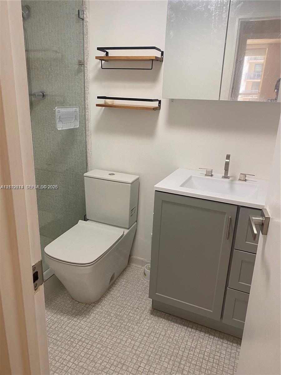 Bathroom featuring a gray vanity cabinet with an integrated white sink, wall-mounted mirrored medicine cabinet, glass-enclosed shower, dual tiered wood and metal wall shelving, and tile flooring Bathroom featuring a gray vanity cabinet with an integrated white sink, wall-mounted mirrored medicine cabinet, glass-enclosed shower, dual tiered wood and metal wall shelving, and tile flooring