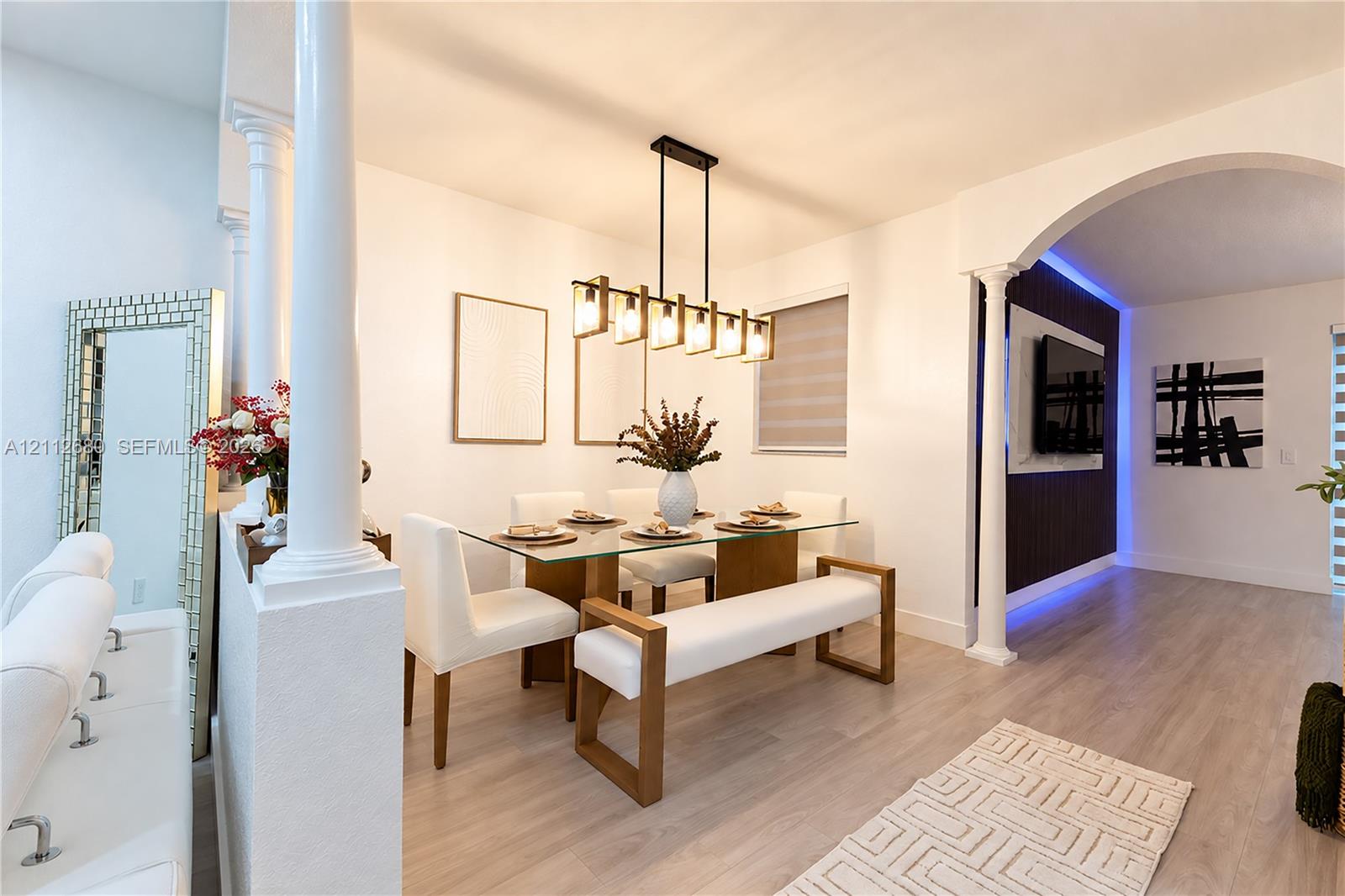 Open-concept area featuring decorative columns, arched architectural detail, linear multi-light pendant fixture, wood-finish flooring, and a recessed accent wall with integrated perimeter lighting