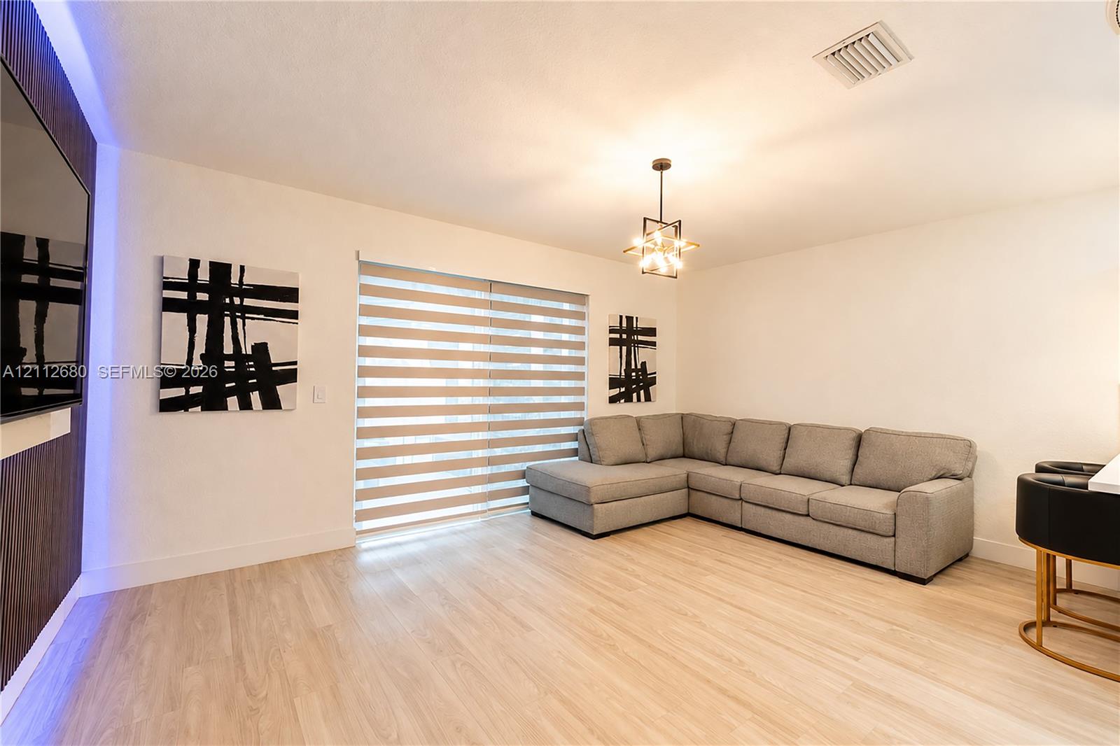 Living area featuring wood-finish flooring, a contemporary multi-light pendant fixture, integrated blue accent lighting, and dual-layered window shades