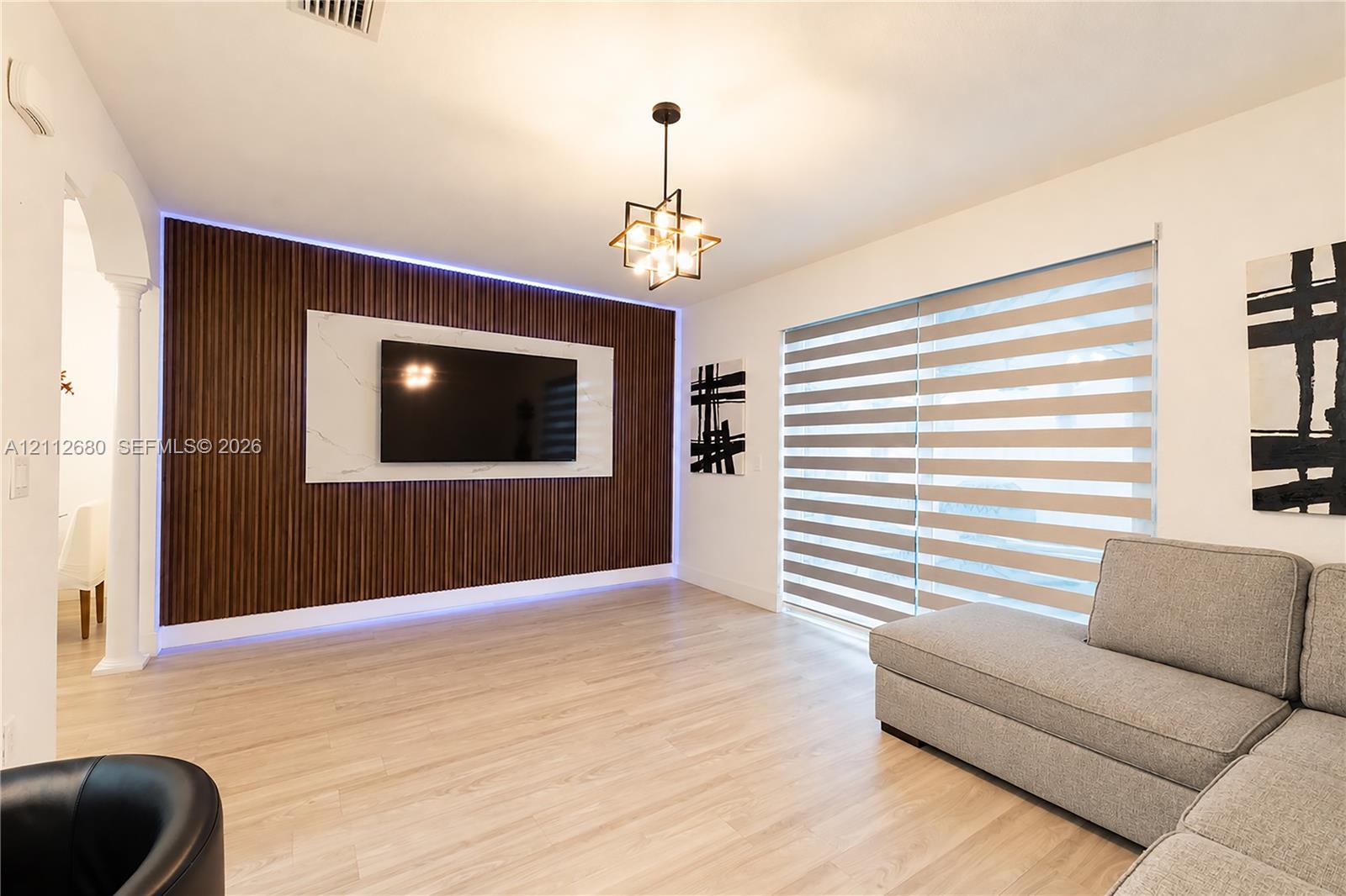 Vertical wood-slat accent wall with integrated LED lighting, stone-finish media panel, wood-finish flooring, geometric multi-bulb chandelier, and large dual-pane horizontal blind window covering Vertical wood-slat accent wall with integrated LED lighting, stone-finish media panel, wood-finish flooring, geometric multi-bulb chandelier, and large dual-pane horizontal blind window covering