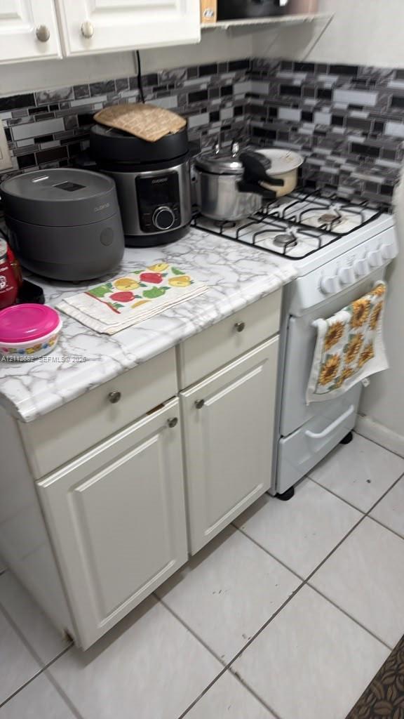 Kitchen featuring white shaker-style cabinetry, a stone-pattern laminate countertop, a four-burner gas range, a multi-tonal glass tile backsplash, and tile flooring