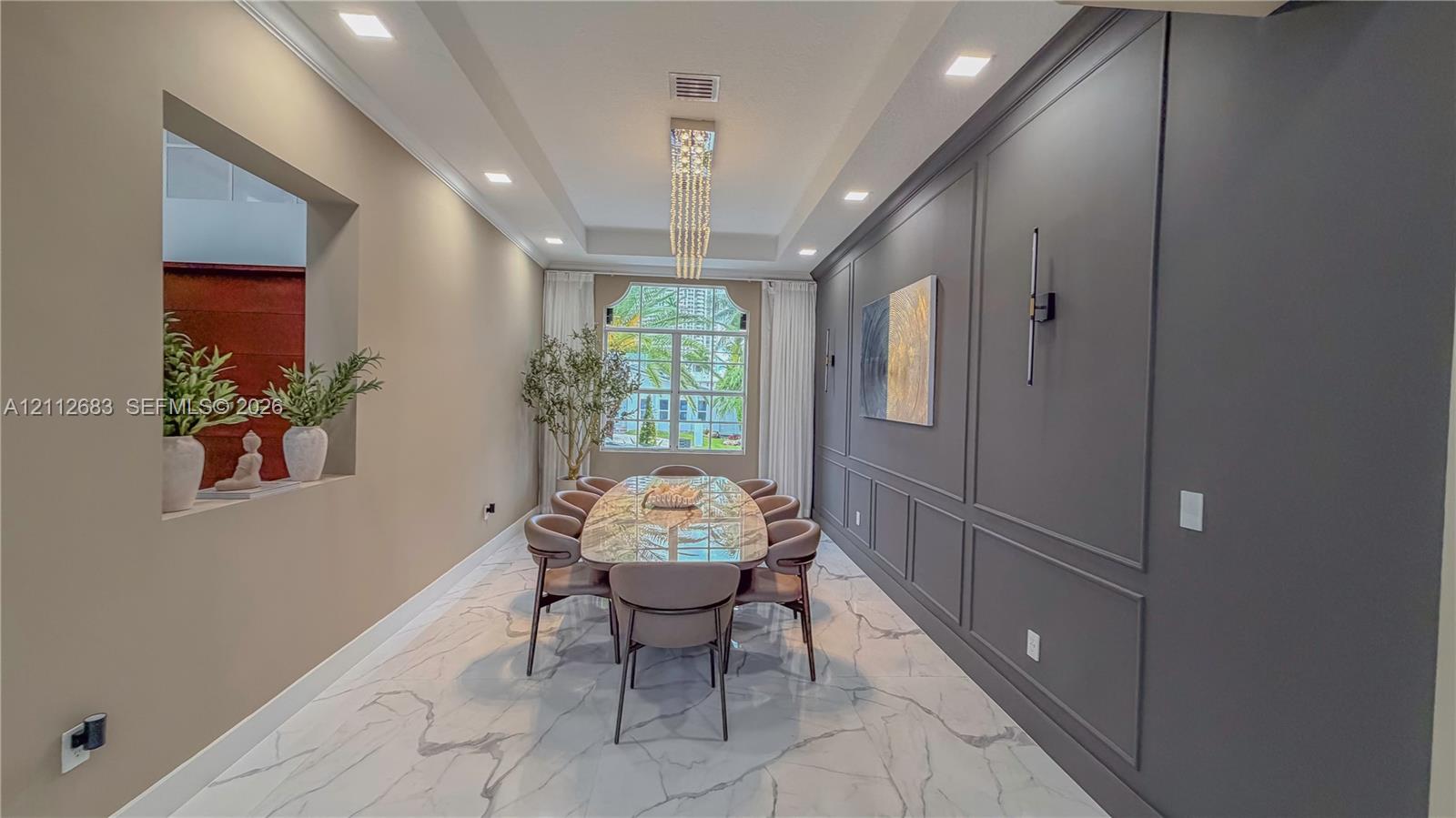 Dining area featuring large-format tile flooring, a tray ceiling with a linear crystal chandelier, an accent wall with wainscoting, and a recessed architectural wall niche