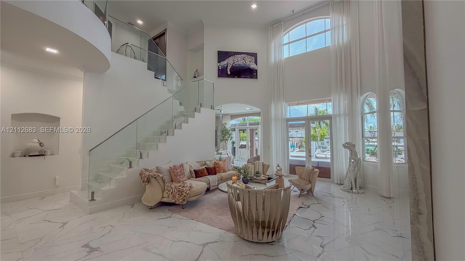 Grand foyer featuring high ceilings, large-format stone flooring, a sweeping staircase with a glass panel railing, arched windows, and a built-in recessed wall niche
