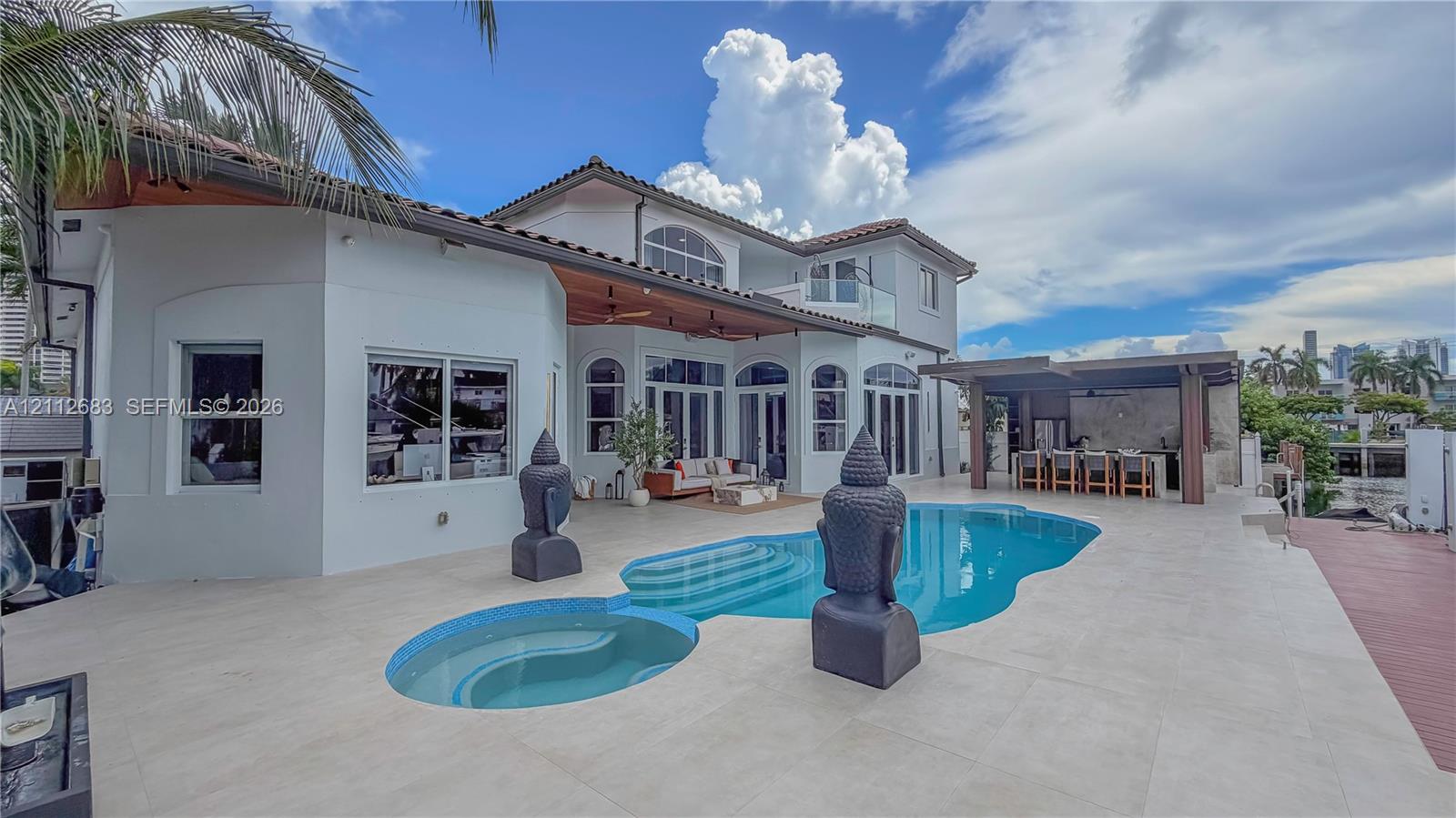 Expansive stone-paved patio featuring a custom-shaped swimming pool, integrated spa, covered outdoor kitchen area, and recessed ceiling lighting