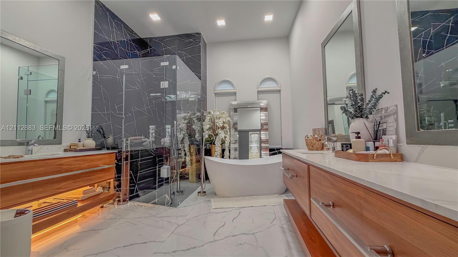 Dual floating wood-finish vanities with light-toned stone countertops, a freestanding soaking tub, a glass-enclosed shower with dark veined wall tile, and recessed lighting