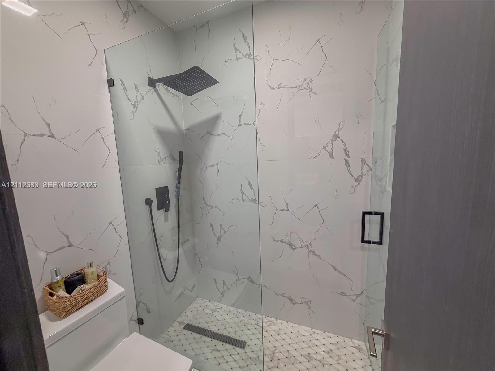 Walk-in shower featuring floor-to-ceiling stone-finish tile, frameless glass enclosure, matte black rain-style shower head with handheld attachment, and recessed wall niche
