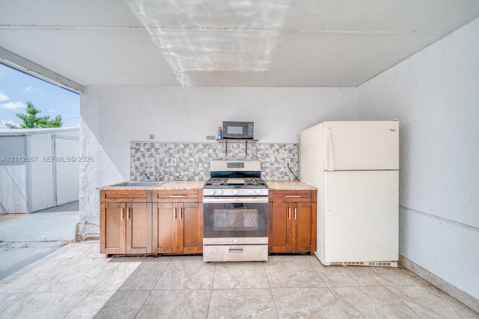 Outdoor kitchen featuring wood-finish cabinetry, stone-look countertop, tiled backsplash, stainless steel gas range, and refrigerator
