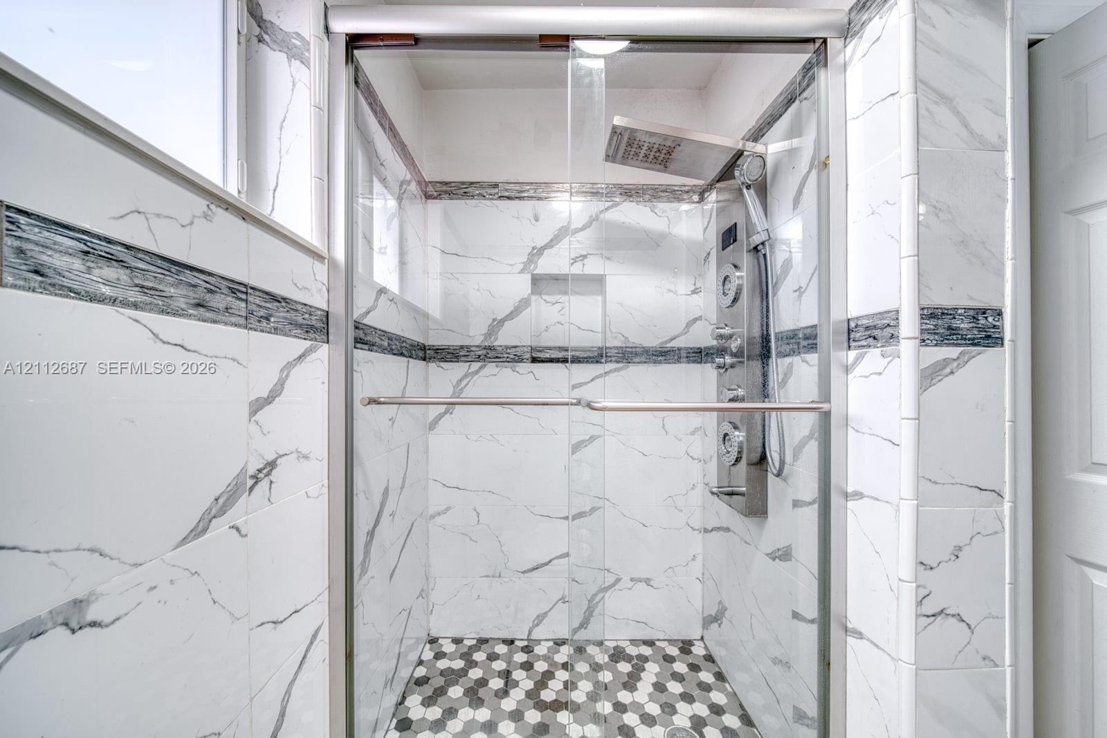 Walk-in shower with floor-to-ceiling veined tile, integrated recessed shelving, multi-head rainfall shower panel, and hexagonal mosaic tile flooring