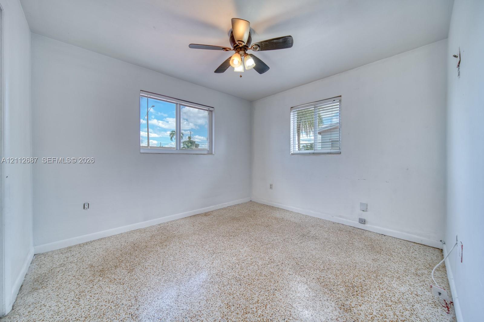Neutral-toned terrazzo flooring, two windows with horizontal blinds, and a central ceiling fan with multi-light fixture
