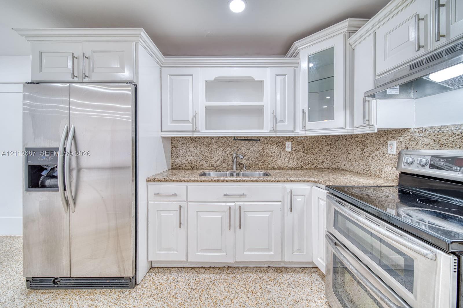Kitchen featuring white shaker-style cabinetry, stone-finish countertops, stainless steel appliances, glass-front upper cabinet, and double-basin undermount sink