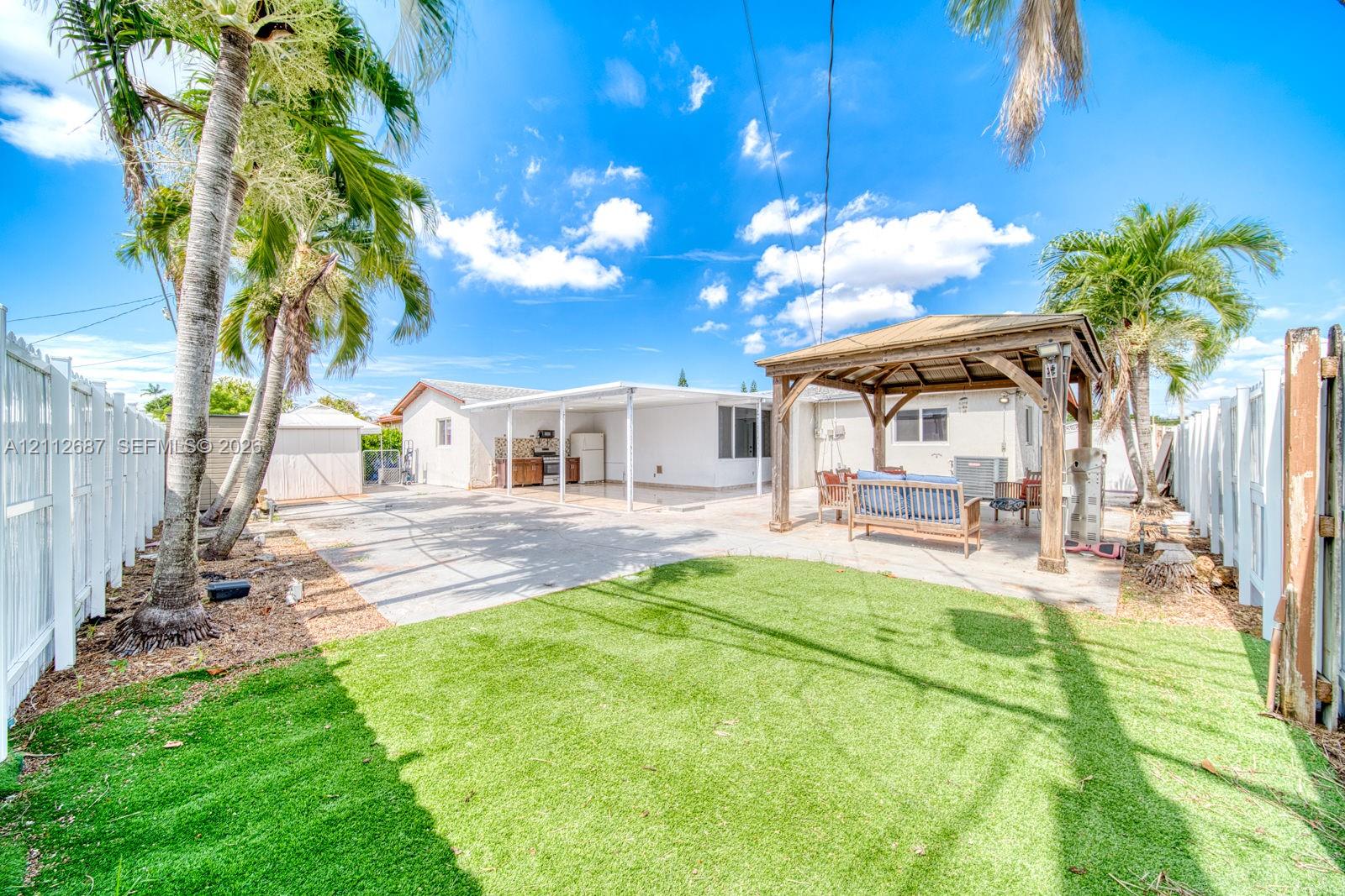 Fenced backyard featuring synthetic turf, a covered concrete patio, a wooden gazebo, and mature palm trees