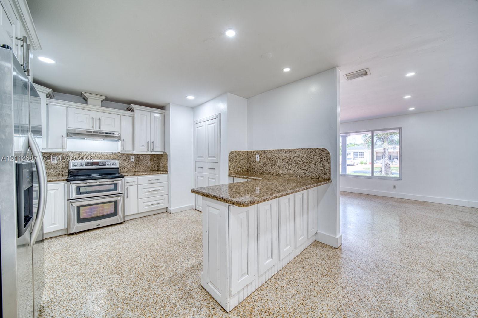 Kitchen featuring white shaker-style cabinetry, stone-surface countertops, stainless steel appliances, a peninsula with bar seating, and polished aggregate flooring