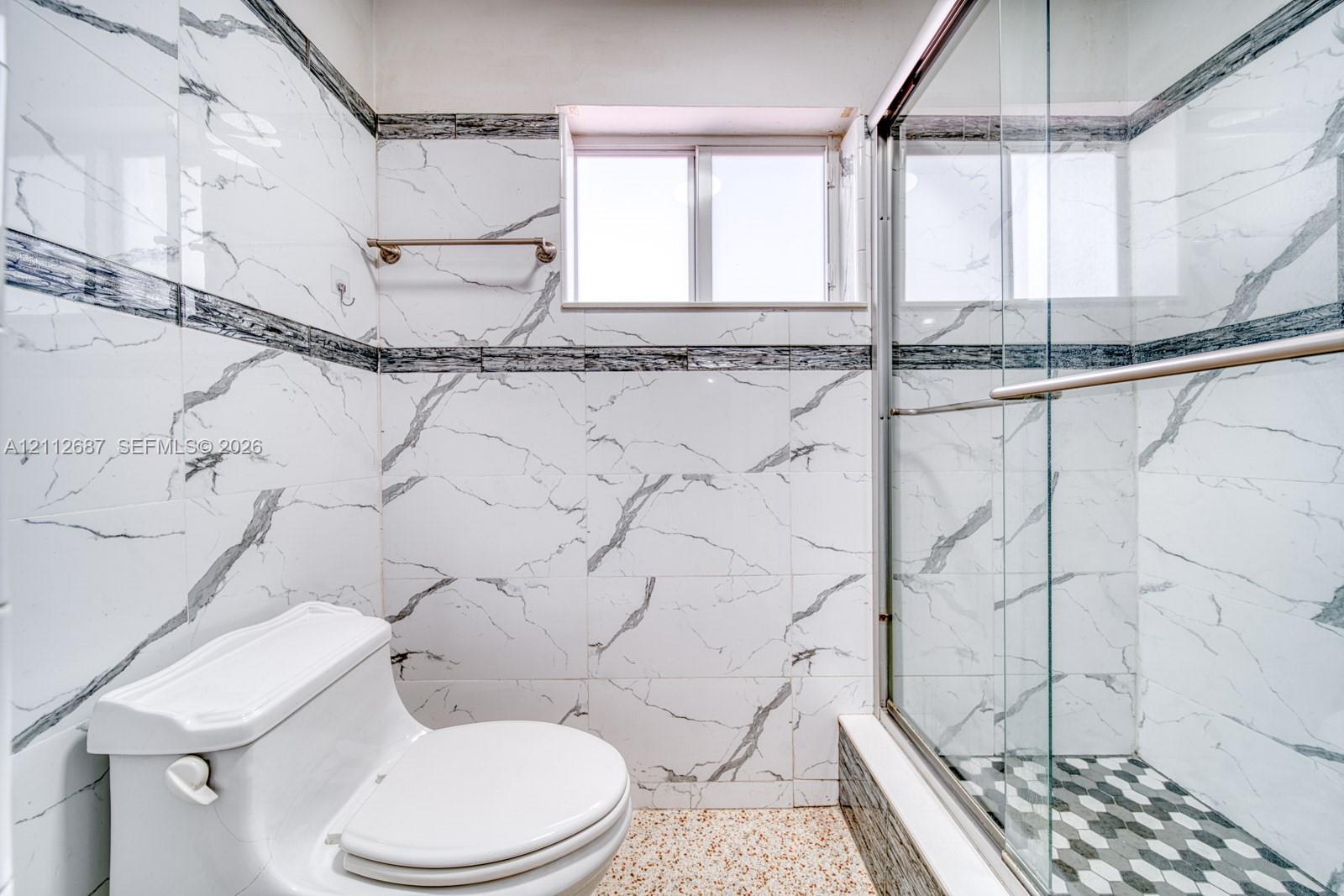 Bathroom featuring floor-to-ceiling white veined wall tile, glass-enclosed shower with hexagonal tile flooring, and a sliding window