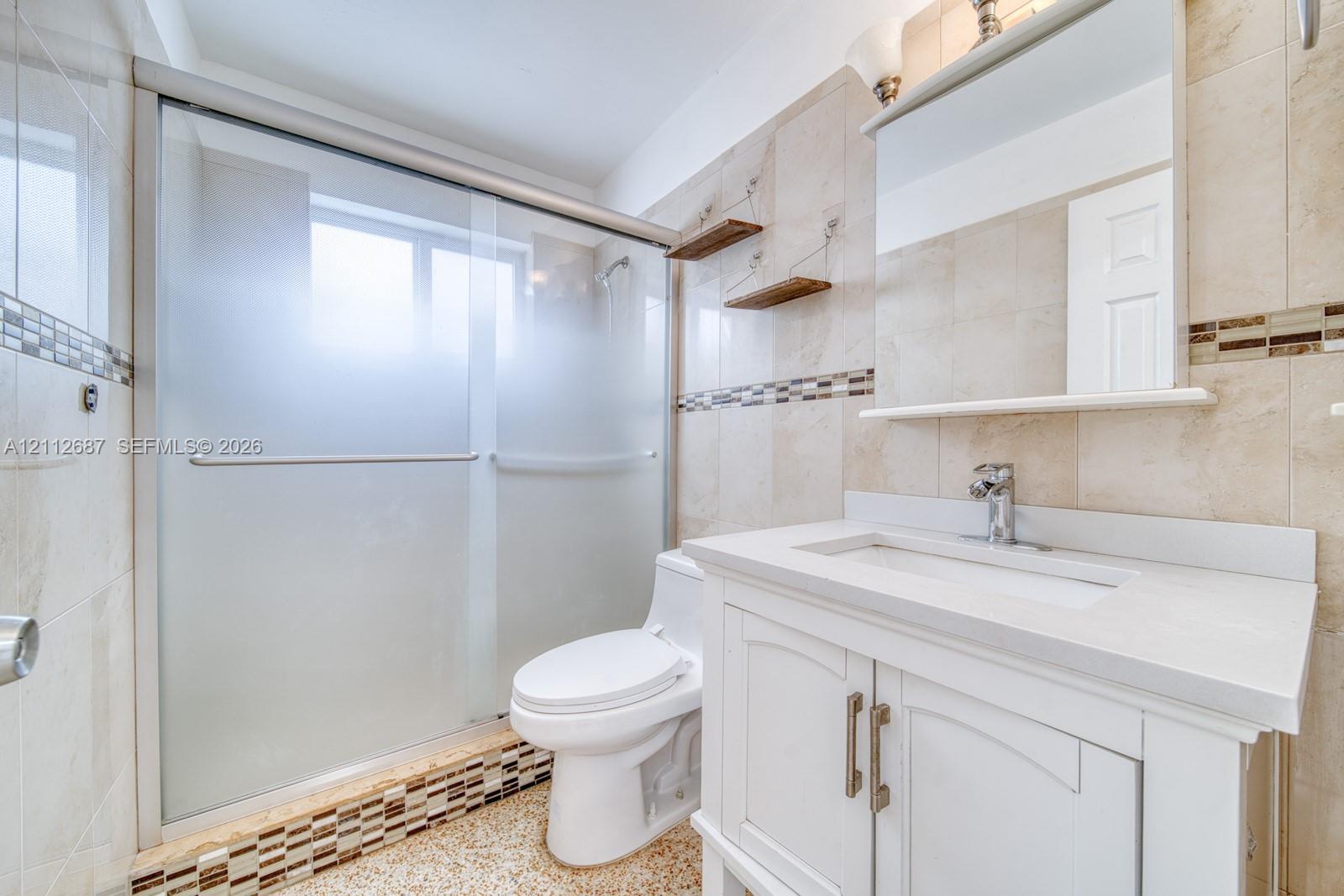 Bathroom featuring a white vanity with a composite countertop, tiled shower enclosure with sliding frosted glass doors, stone-tiled walls with mosaic inlay, and wall-mounted shelving