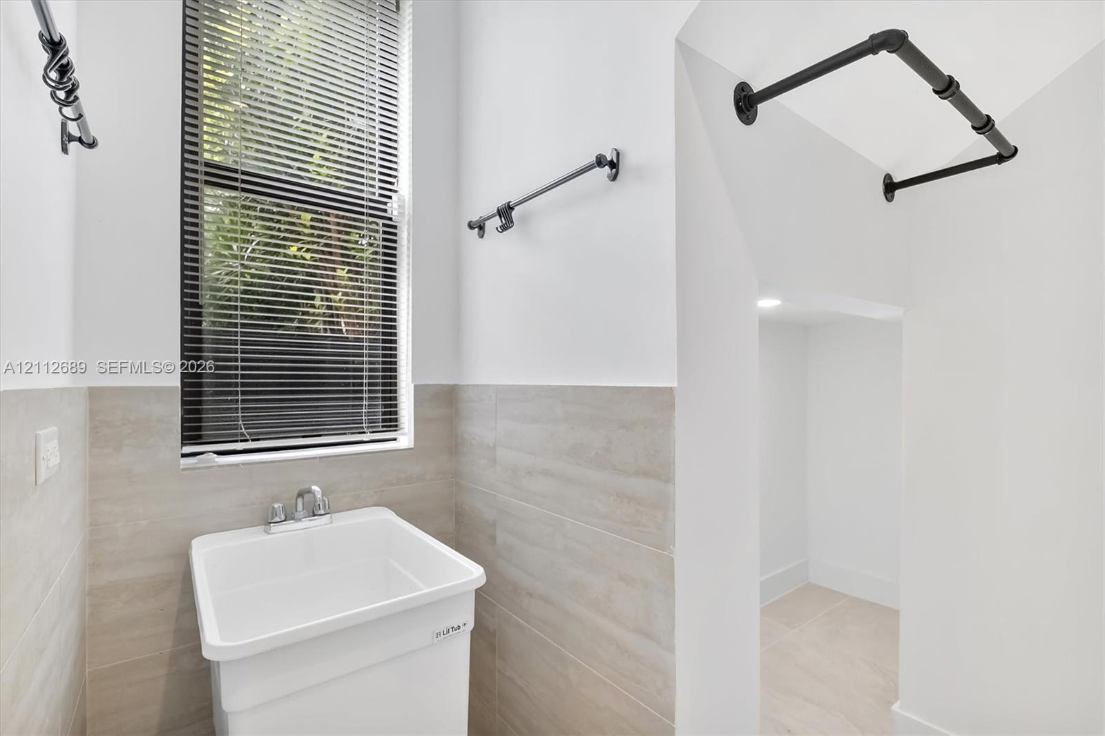Utility space featuring a basin sink with chrome faucet, neutral wall-tile wainscoting, matte black wall-mounted hardware, and an open storage alcove
