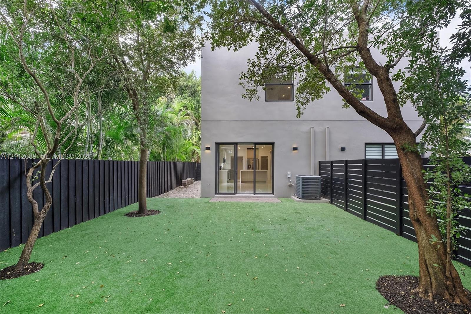 Fenced backyard featuring synthetic turf, mature trees, smooth stucco exterior, and sliding glass doors