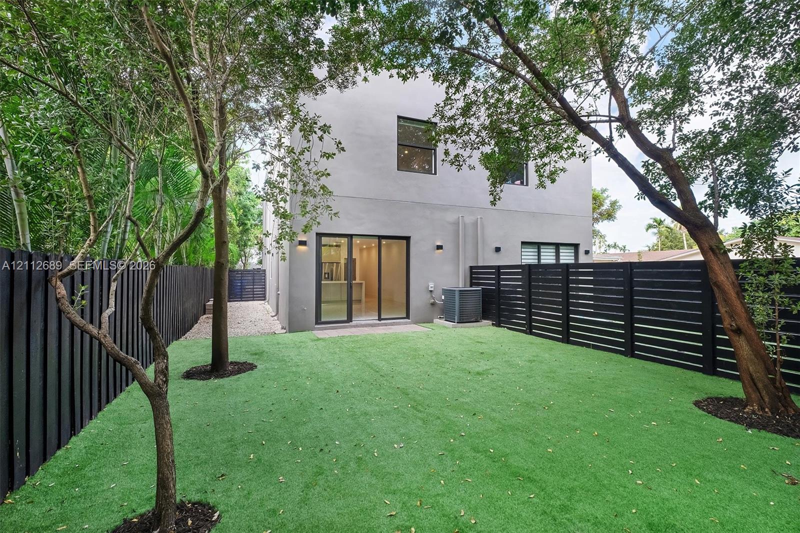 Fenced backyard featuring synthetic turf, mature trees, smooth stucco exterior, and sliding glass doors