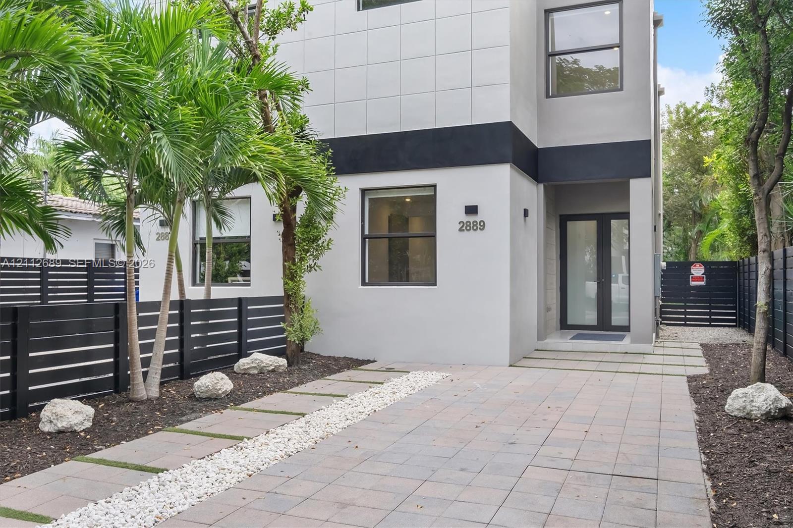 Modern two-story exterior featuring smooth stucco siding, large rectangular windows, black horizontal perimeter fencing, paver driveway, and a glass-paneled entry door