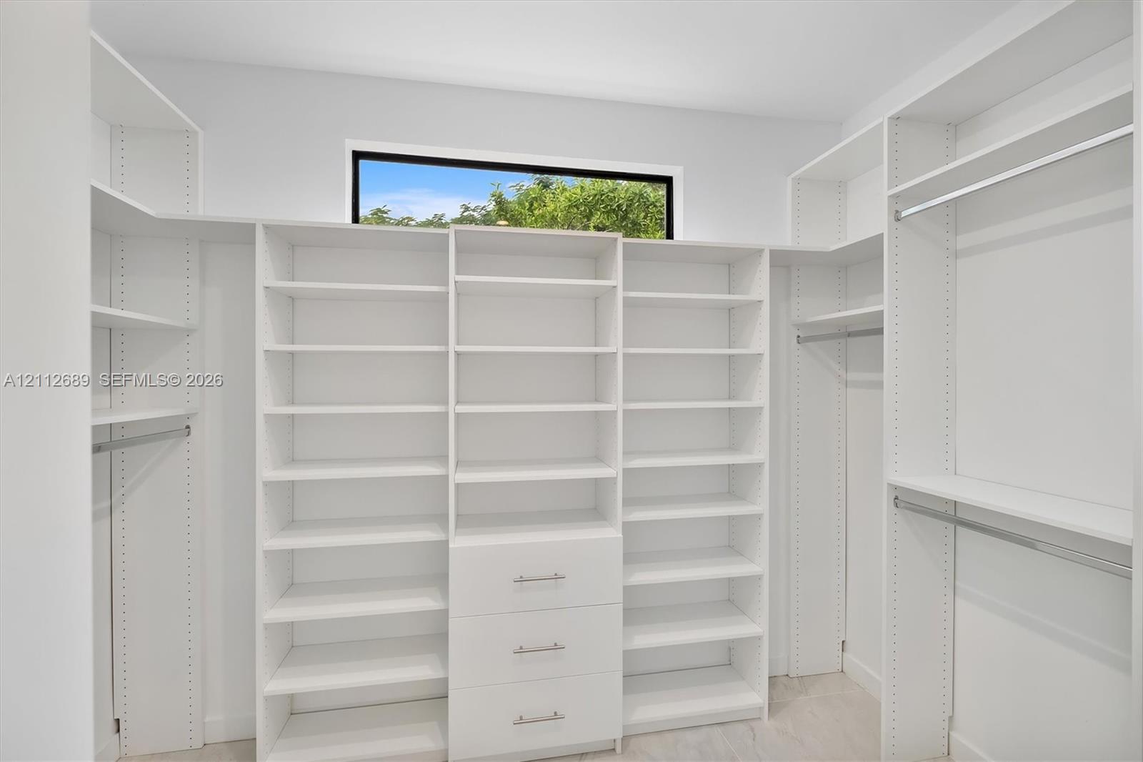 Walk-in closet featuring floor-to-ceiling white built-in shelving, integrated dresser drawers with metallic pulls, horizontal hanging rods, and a clerestory window