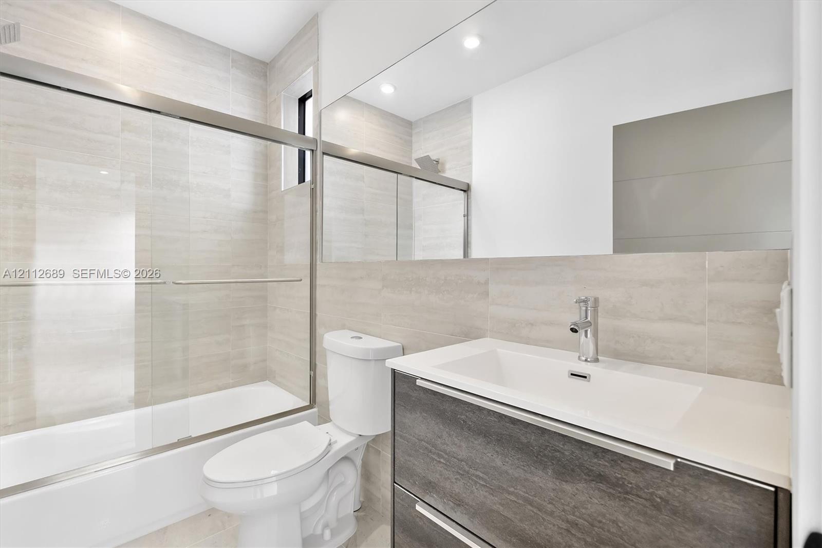 Bathroom featuring a wall-to-wall mirror, modern floating vanity with a stone-top integrated sink, floor-to-ceiling tile surround, and a glass-enclosed tub shower with metallic hardware