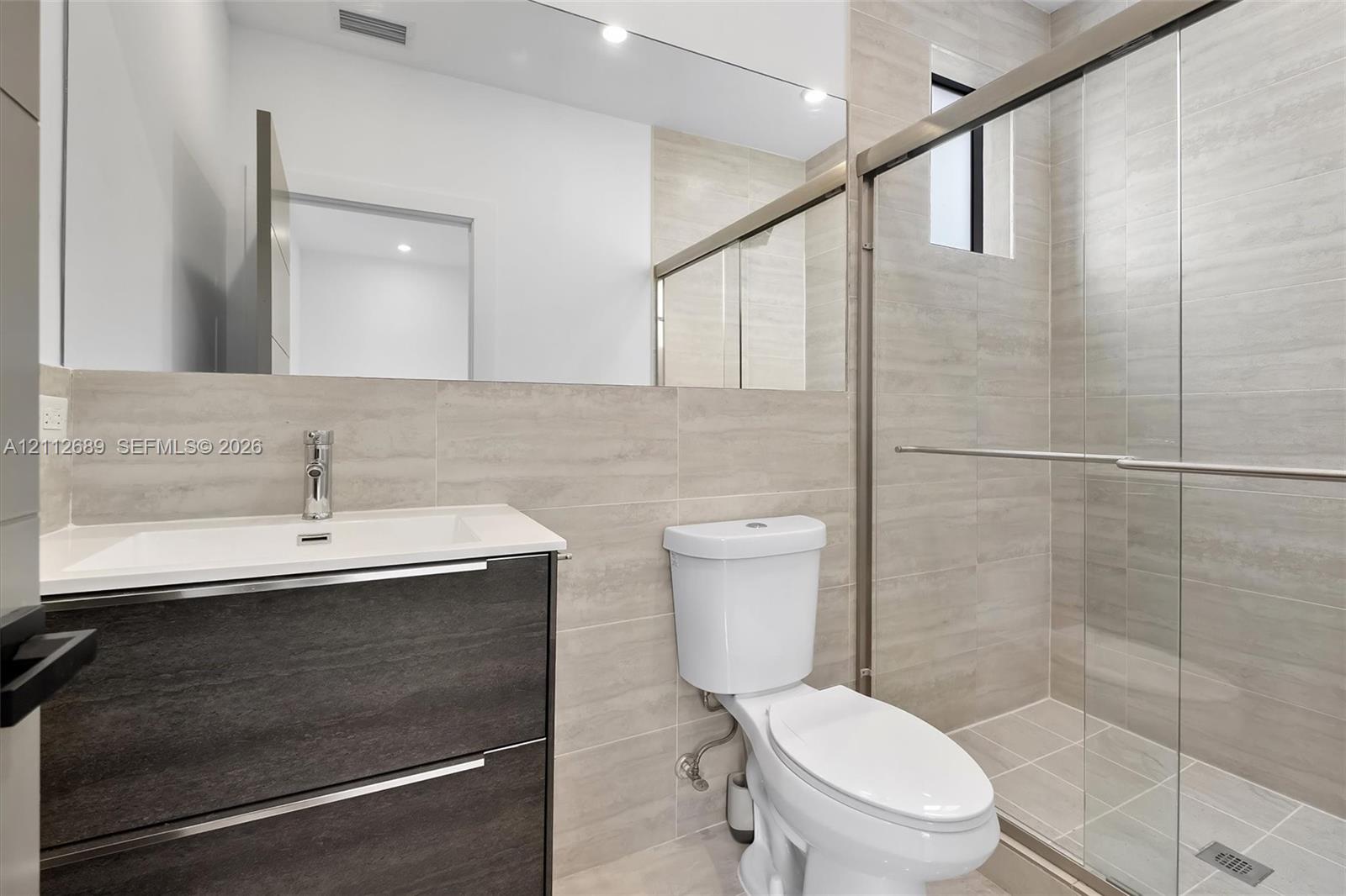 Wall-mounted vanity with a rectangular sink, full-width mirror, walk-in shower with sliding glass doors, and neutral large-format tile wall cladding