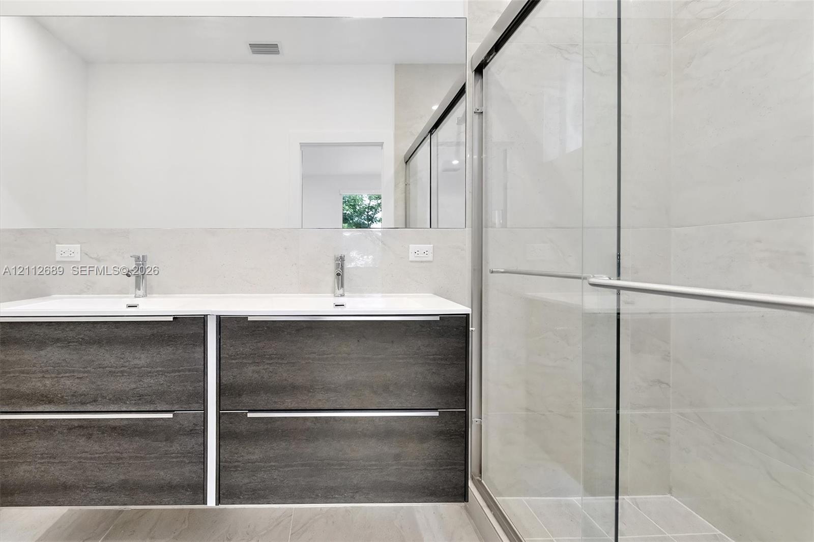 Floating wood-finish double vanity with dual undermount sinks and chrome fixtures, large wall-mounted mirror, and walk-in shower featuring glass sliding doors and floor-to-ceiling tile surround
