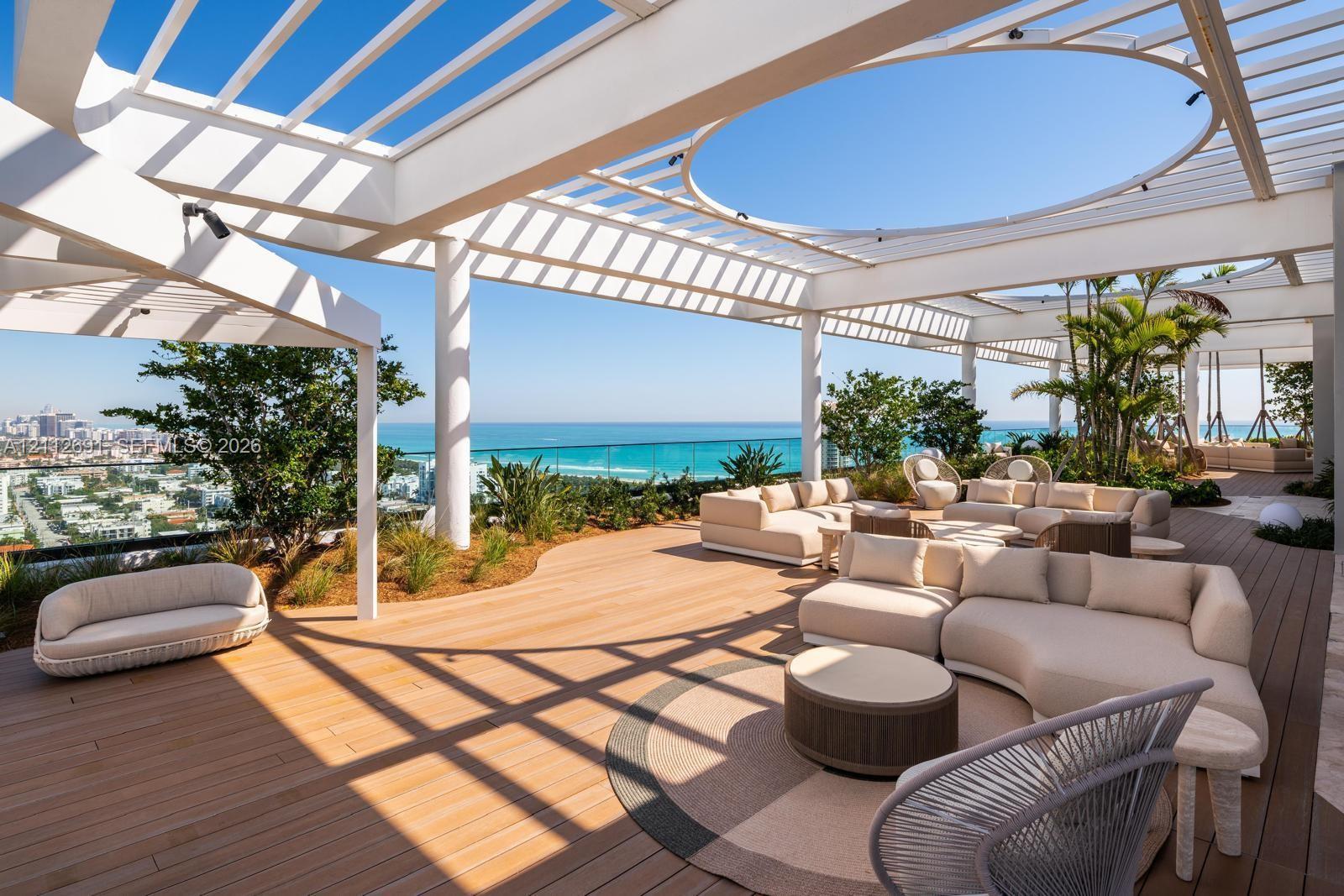 Expansive rooftop terrace featuring a white pergola structure with a circular cutout, wood-finish decking, glass panel railings, and perimeter landscaping