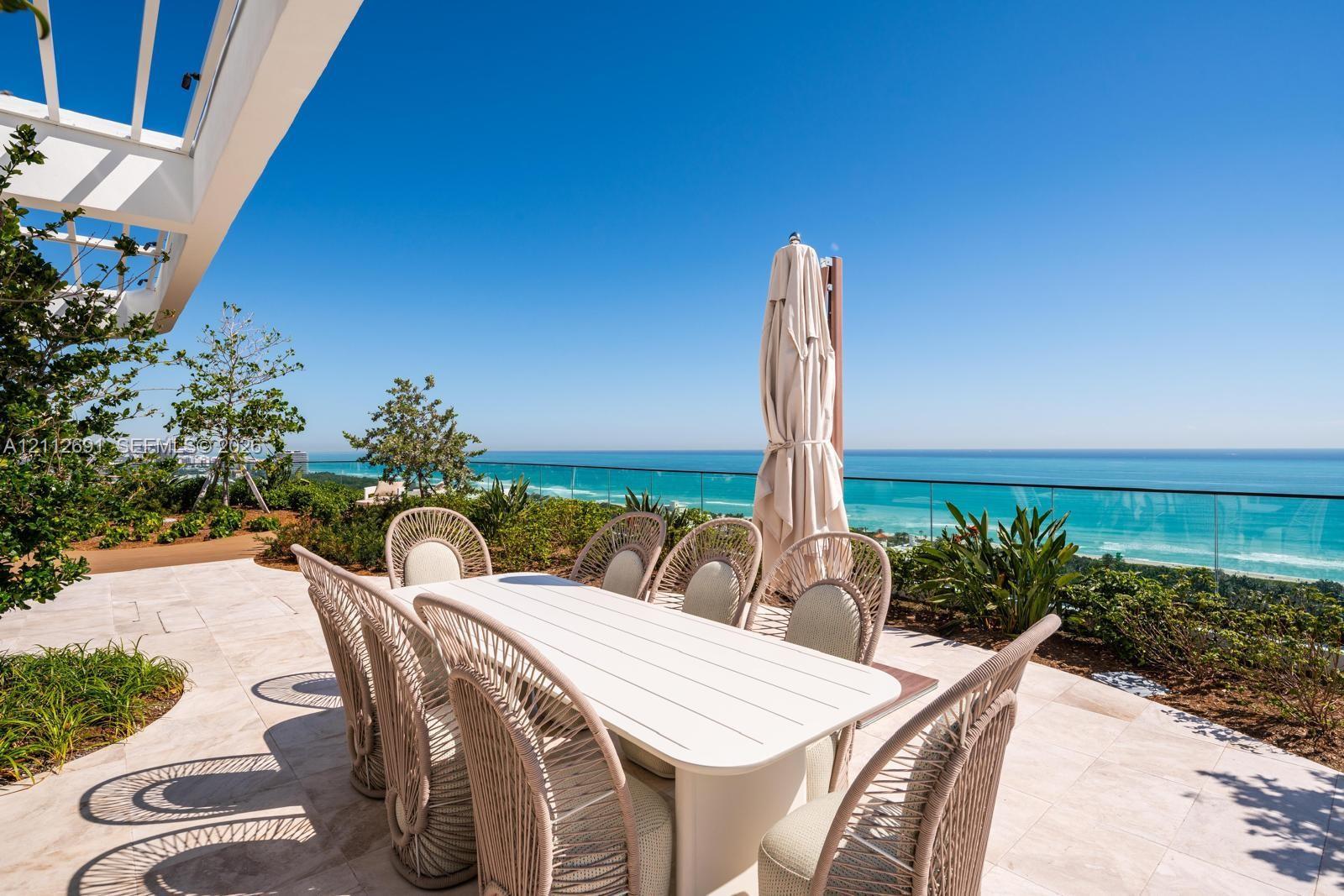 Expansive stone-paved terrace featuring a white pergola, transparent glass railing, and mature coastal landscaping