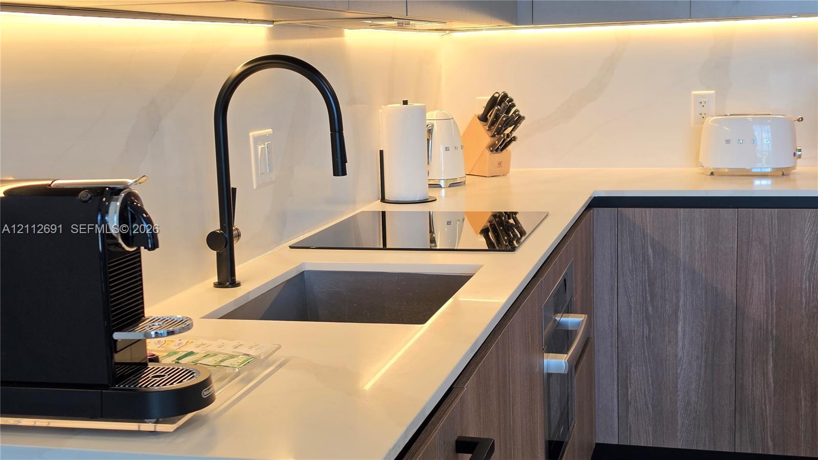Kitchen featuring light-toned countertops, integrated under-cabinet lighting, wood-finish cabinetry, a square undermount sink, and a matte black high-arc faucet