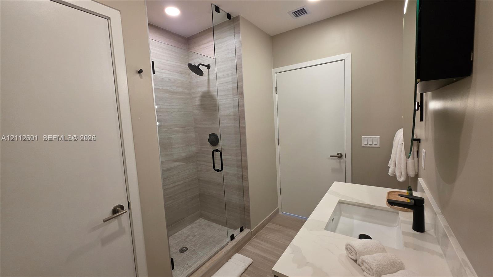 Bathroom featuring a walk-in shower with frameless glass enclosure, horizontal stone-finish tile surround, recessed lighting, white vanity with stone-finish countertop, and matte black plumbing fixtures