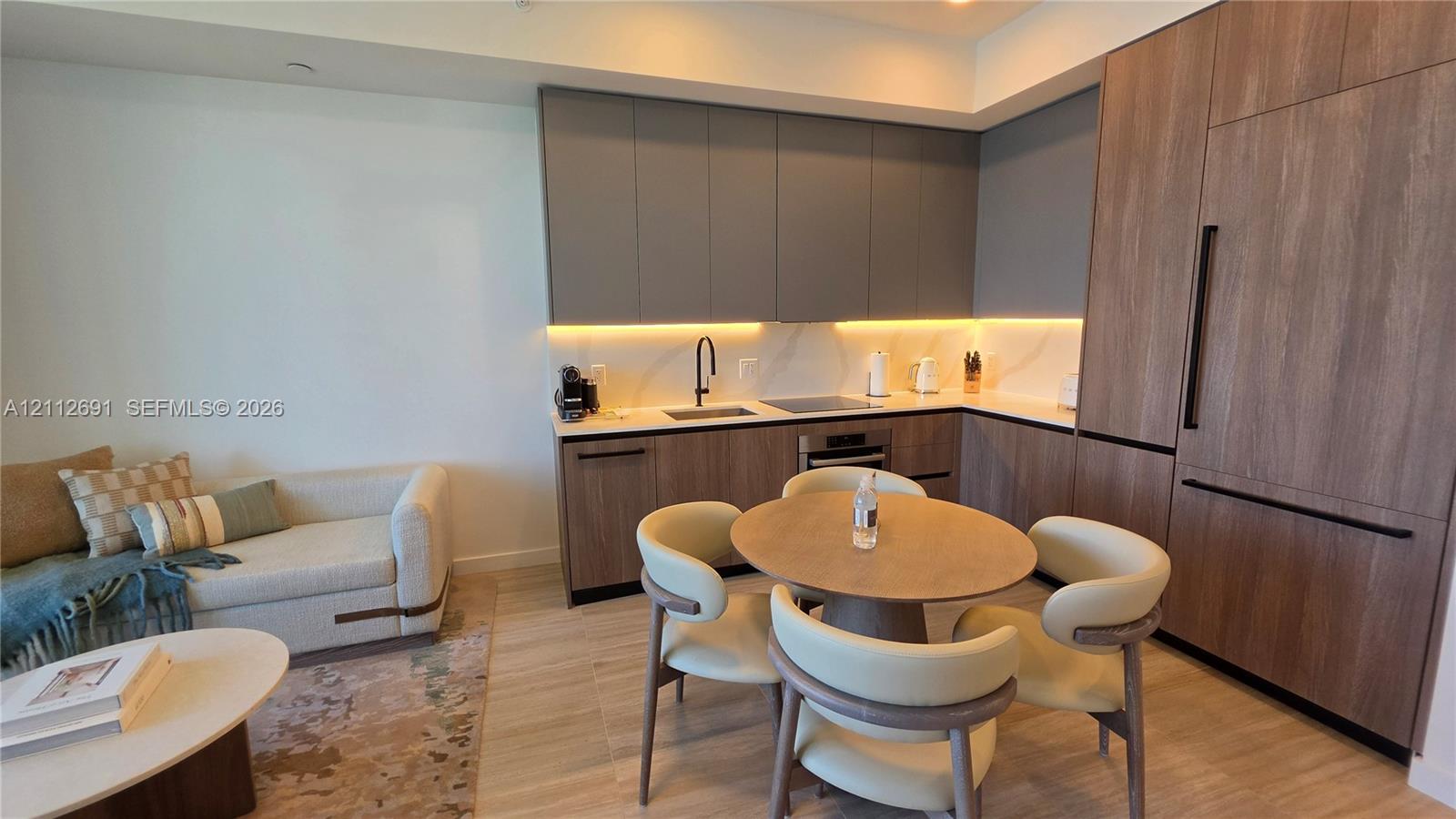 Kitchen featuring flat-panel cabinetry in matte grey and wood-finish, light-toned stone-finish countertops, integrated refrigerator, and under-cabinet lighting