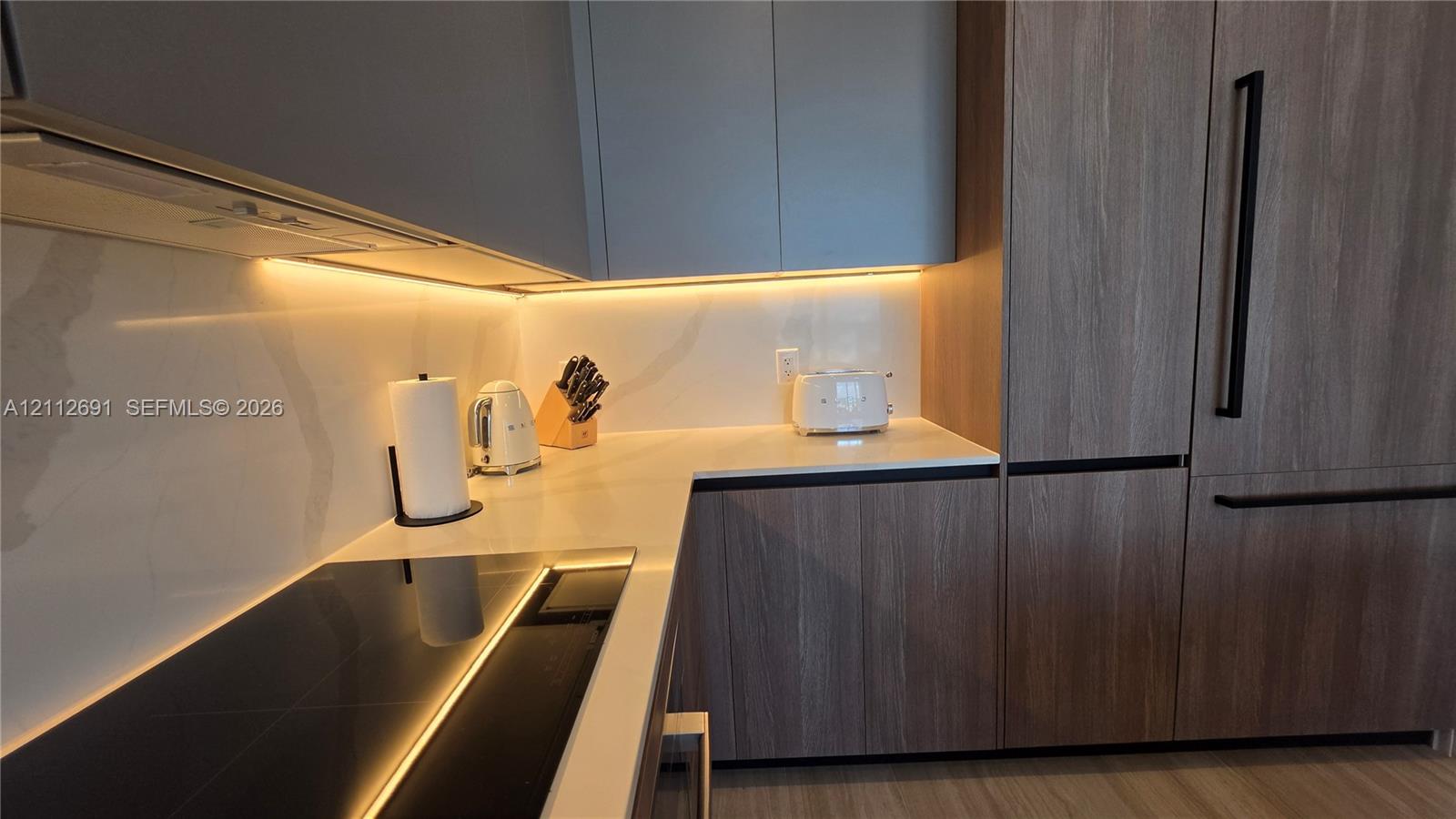Modern kitchen featuring flat-panel cabinetry with wood-finish grain, integrated paneled refrigerator, solid-surface backsplash, under-cabinet lighting, and induction cooktop