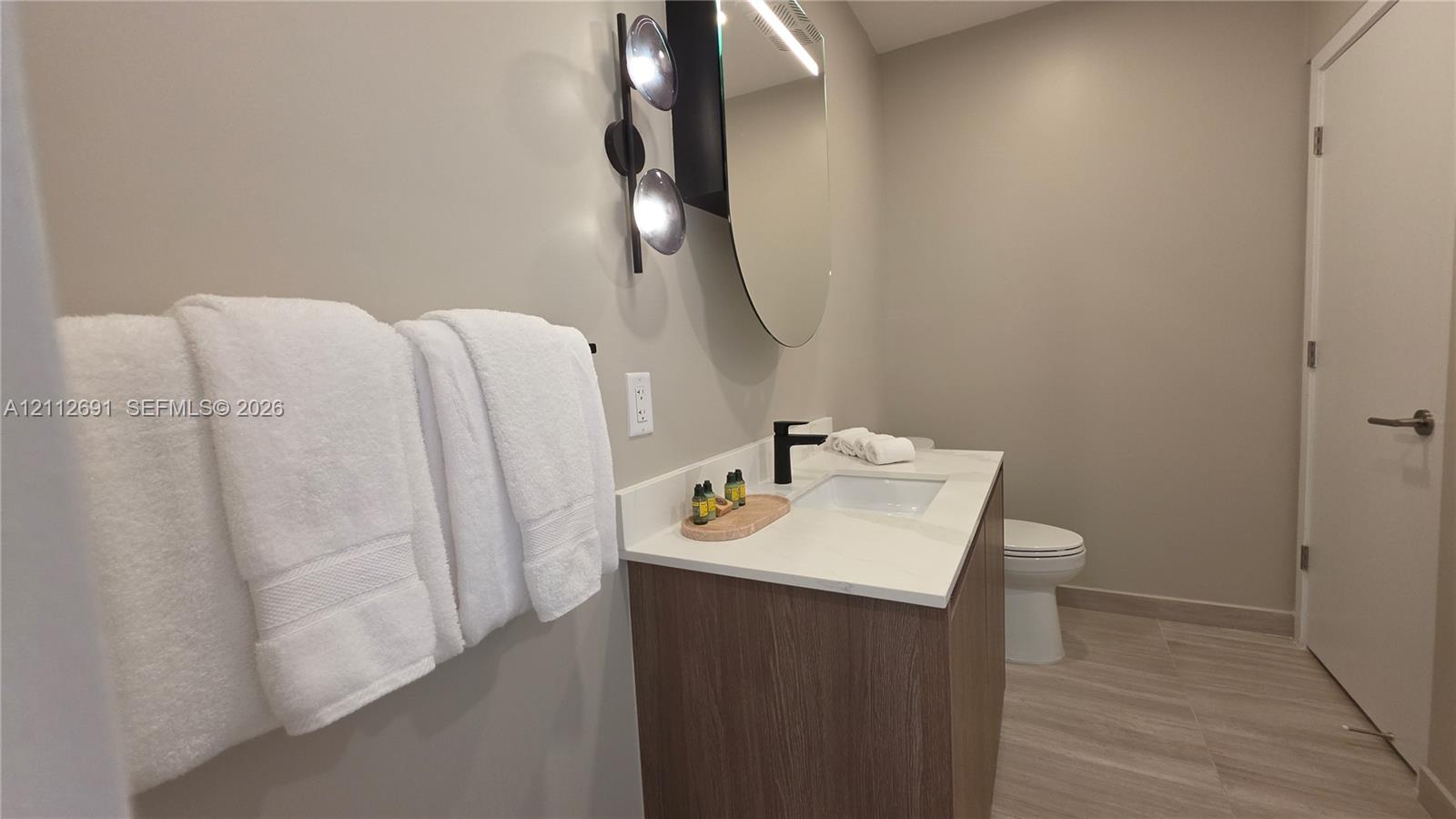 Modern vanity with wood-finish cabinetry, white countertop, integrated rectangular sink, matte black faucet, and oval mirror with wall-mounted lighting