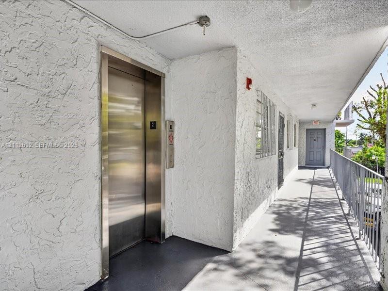 Exterior walkway featuring a brushed-metal elevator door, textured stucco wall finishes, and a metal-railing enclosure