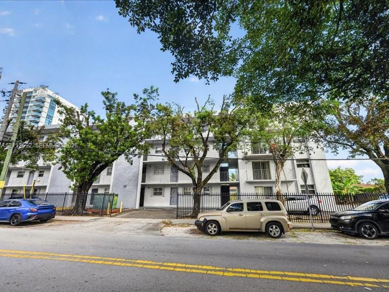 Multi-story residential building exterior featuring a light-toned stucco facade, private balconies, and perimeter metal fencing