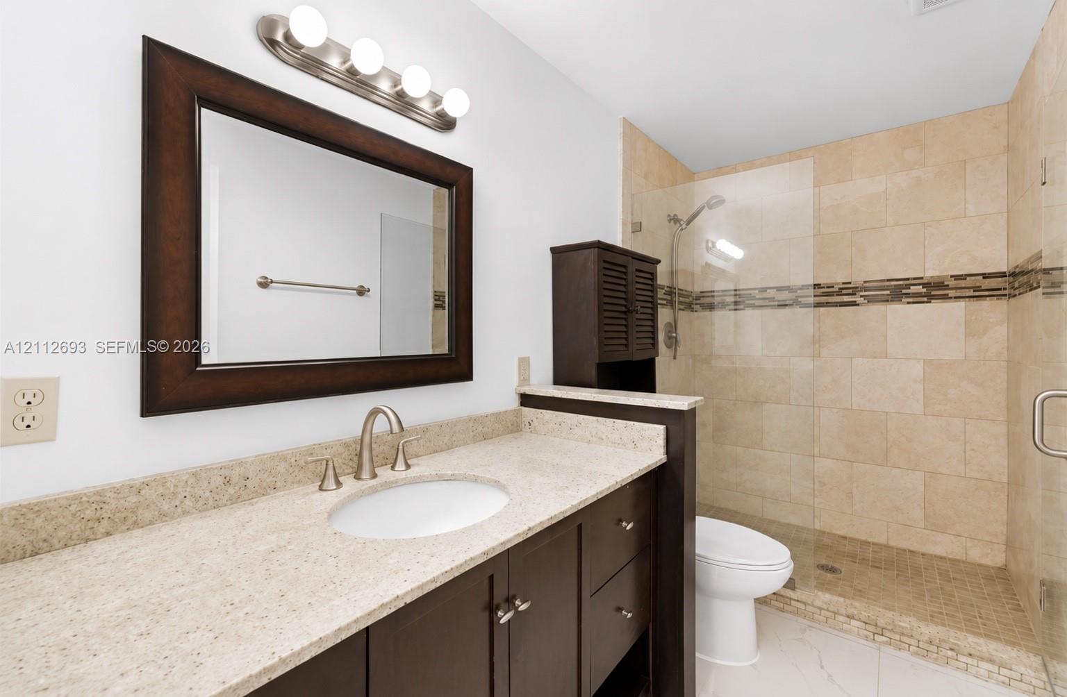 Bathroom featuring a dark-finish wood vanity with a stone countertop, an integrated oval sink, a glass-enclosed walk-in shower with stone tile surround, and a coordinating vanity mirror with an attached four-light fixture