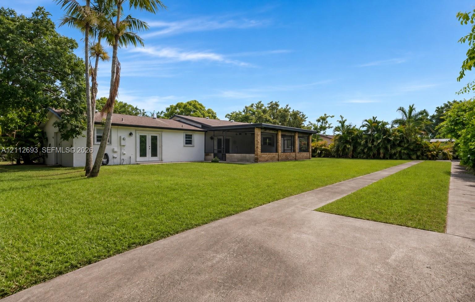 Expansive backyard featuring a sprawling lawn, mature palm trees, a concrete driveway, and a covered patio area with masonry detailing