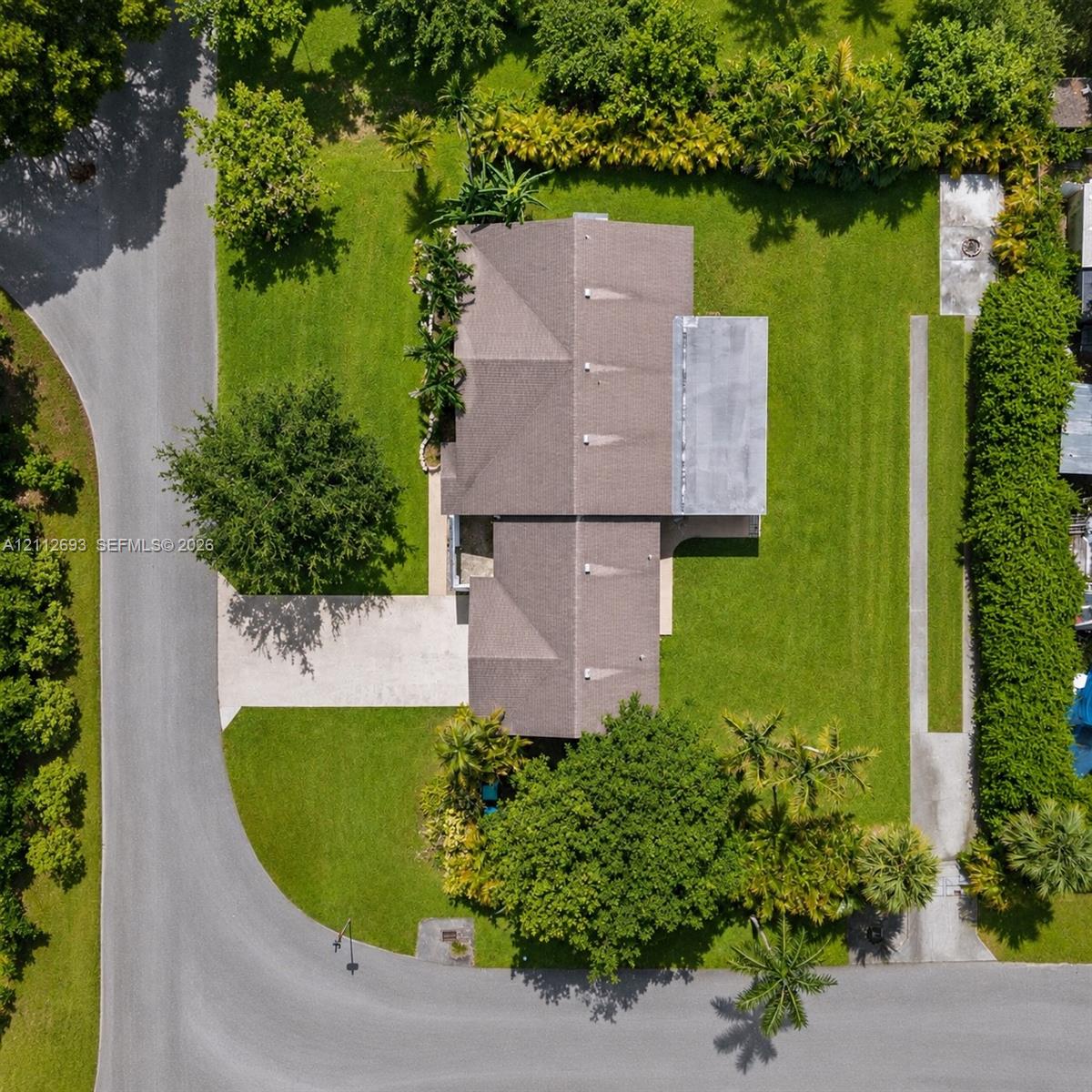 Aerial perspective featuring a multi-hipped roofline, expansive grass lawn, paved driveway, mature trees, and dense perimeter landscaping