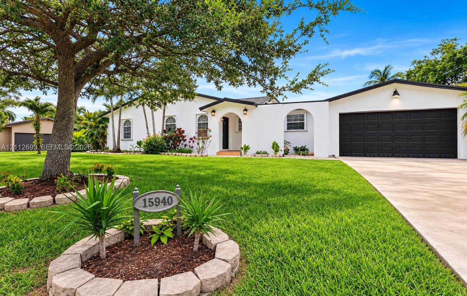 Single-story exterior featuring a crisp white facade, arched entryway, dark-toned multi-car garage door, concrete driveway, and stone-bordered landscaping