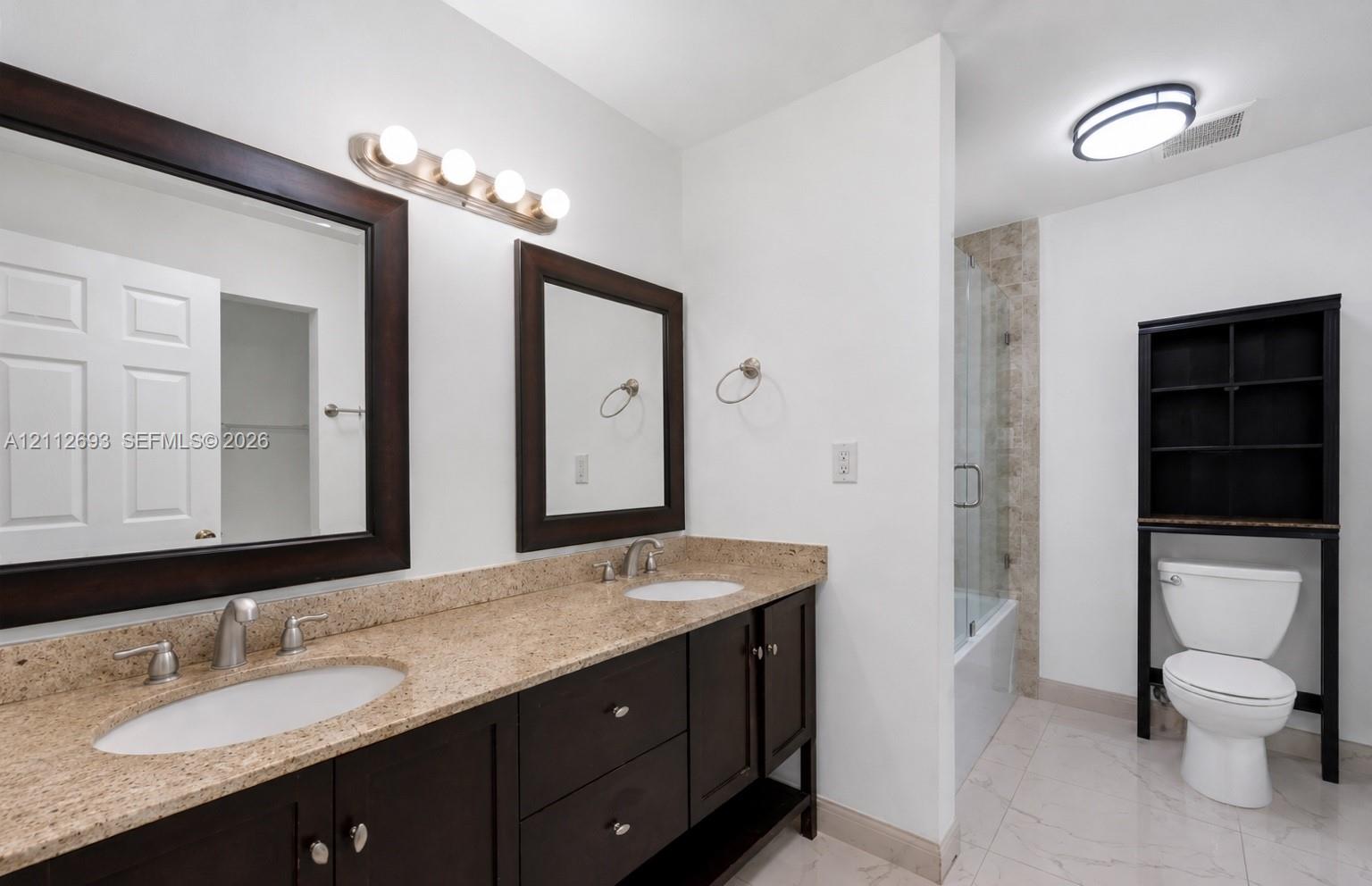 Dual-sink vanity with stone countertop, dark cabinetry, framed mirrors, and a glass-enclosed shower featuring tile surrounds