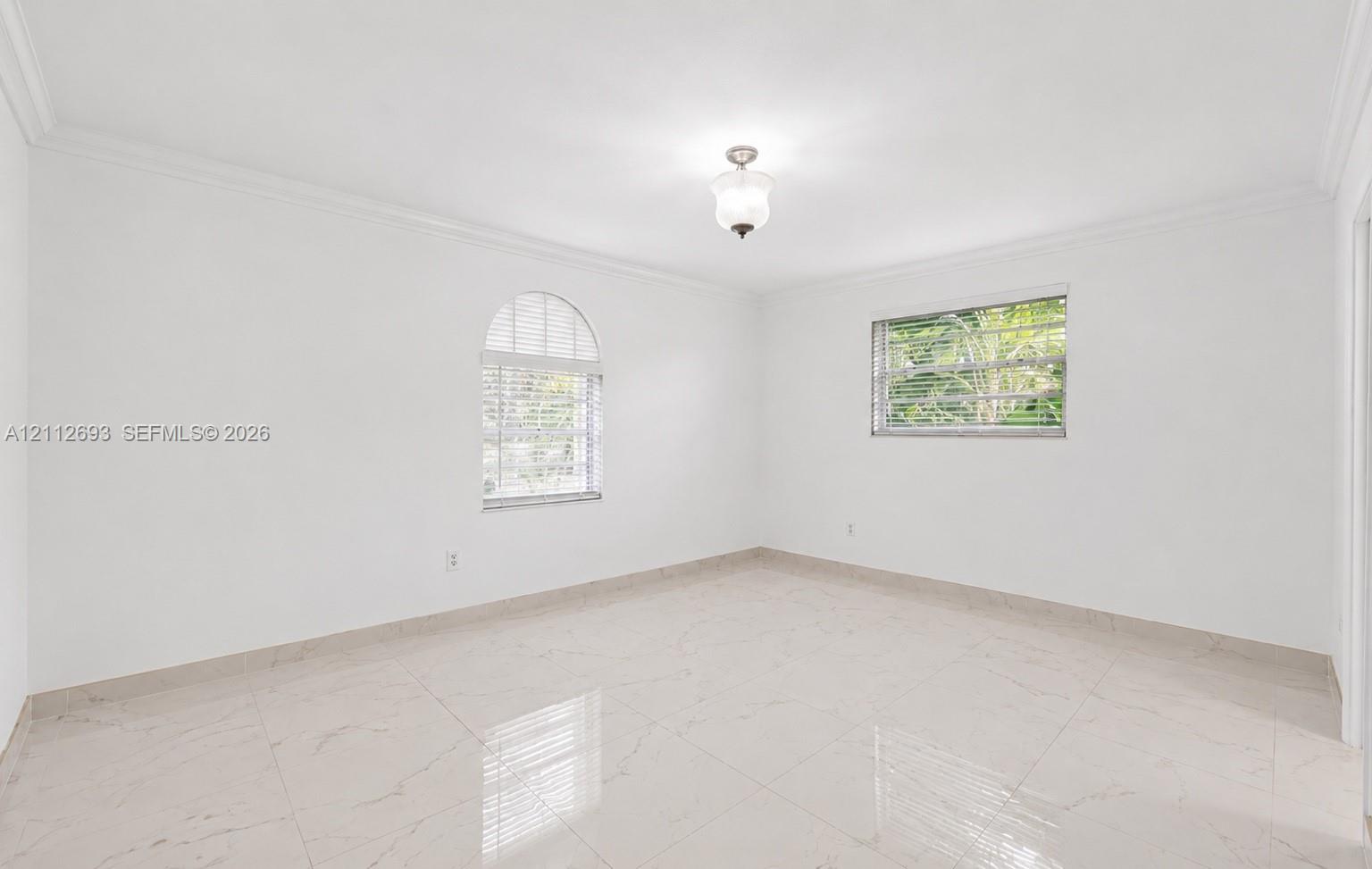 Neutral-toned interior space featuring polished tile flooring, crown molding, an arched window, and a centered flush-mount light fixture