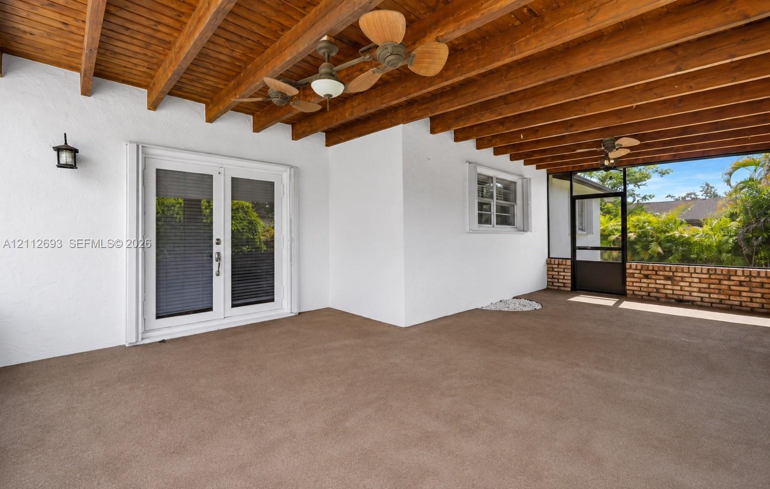 Covered patio featuring exposed wood-beam ceiling, dual ceiling fans, white stucco walls, French door entry, and screened enclosure with brick-accented base