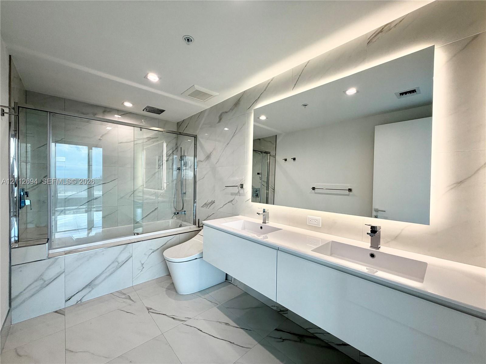 Double floating vanity with integrated rectangular sinks, expansive LED-backlit mirror, large-format stone-finish floor and wall tiles, and glass-enclosed bathtub with chrome fixtures