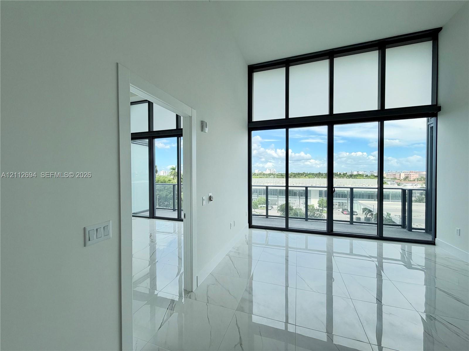 Floor-to-ceiling windows with dark framing, large-format stone-finish tile flooring, and sliding glass doors with access to a railed balcony Floor-to-ceiling windows with dark framing, large-format stone-finish tile flooring, and sliding glass doors with access to a railed balcony